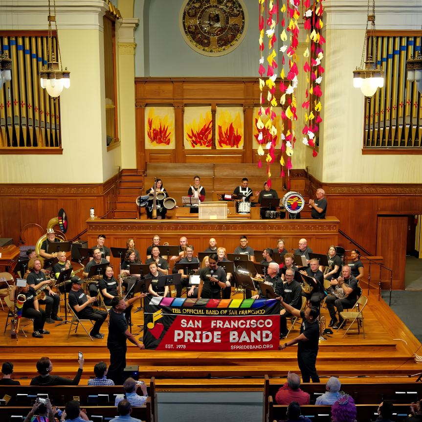San Francisco Pride Band performing on stage, holding their rainbow banner, with an audience in the pews.