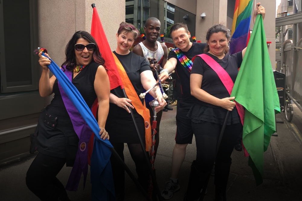 Five Twirling & Flag Corps members pose with rainbow Pride flags on a city sidewalk before the start of the Pride Parade.